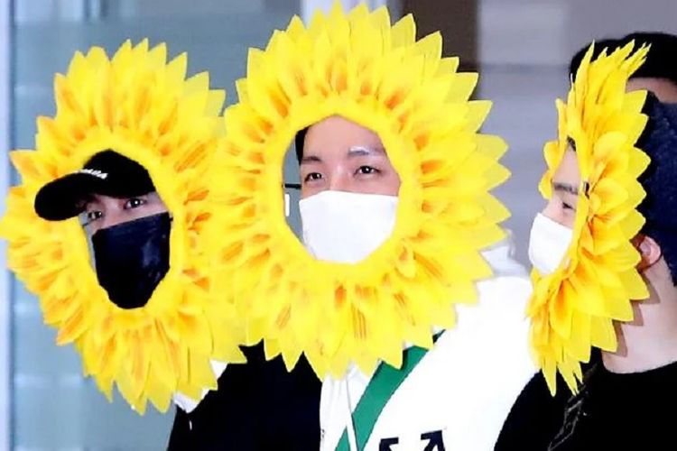 Tiga member BTS, (dari kiri) Jin, J-Hope, dan Suga, memakai hiasan kepala berbentuk kelopak bunga matahari saat tiba di bandara Icheon, Korea Selatan, Senin (10/6/2019).