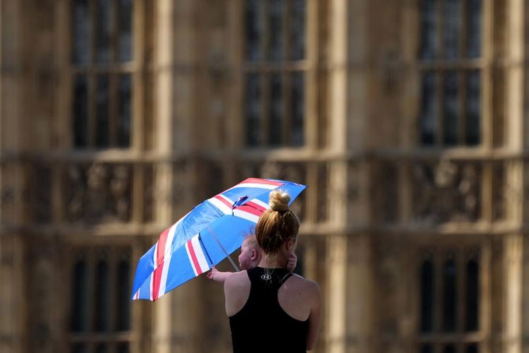 Seorang perempuan menggendong bayi sambil memegang payung untuk berlindung dari suhu ekstrem di Westminster Bridge di London, Inggris, Selasa (19/07/2022).