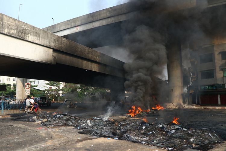 Pengendara sepeda motor melintas menembus kepulan asap sisa barikade yang terbakar di Yangon, pada Senin (15/3/2021).