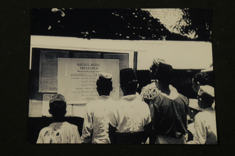 Sekelompok pria berdiri di depan papan yang bertuliskan Romukyokai, kepanitiaan Romusha pada masa pendudukan Jepang.