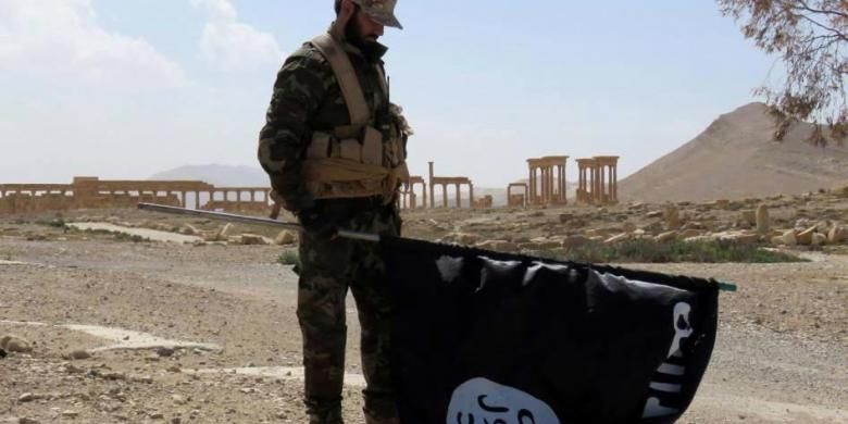 Seorang anggota pasukan pemerintah Suriah memegang bendera ISIS saay berada di jalan kota tua Palmyra yang direbut kembali dari tangan ISIS, Minggu (27/3/2016).