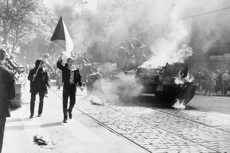 Warga kota Praha membawa bendera Cekoslovakia berjalan melintasi sebuah tank Uni Soviet yang terbakar dalam invasi Pakta Warsawa ke negeri itu pada 20-21 Agustus 1968.