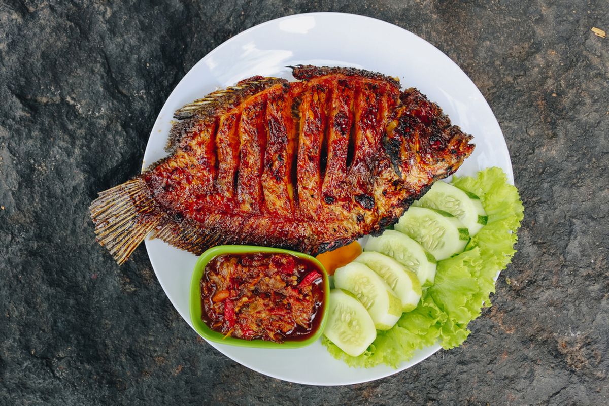 Ilustrasi ikan gurame bakar, sambal, dan lalapan di atas piring. 