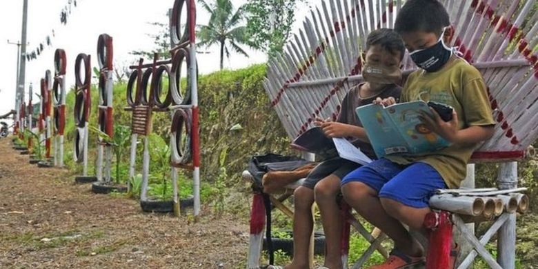 Dua murid sekolah dasar mengikuti proses belajar mengajar secara online di tepi jalan di ruang terbuka.