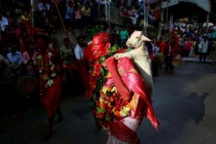 Seorang pendeta Hindu, dengan wajah penuh riasan dan darah hewan kurban, menggendong seekor kambing yang akan dikurbankan dalam festival Deodhani di kuil Kamakhya, Gauhati, India pada 18 Agustus 2014. Dalam festival ini, umat Hindu India mengurbankan kambing dan burung dara.