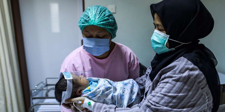 bSeorang ibu menggendong bayinya yang memakai pelindung wajah di Rumah Sakit Ibu dan Anak (RSIA) Tambak, Jakarta Pusat, Rabu (23/5/2020). Rumah Sakit Ibu dan Anak (RSIA) menerapkan kebijakan baru berupa pemakaian face shield atau pelindung wajah untuk bayi yang sedang mendapat perawatan maupun yang baru dilahirkan di RS tersebut. Hal tersebut dilakukan untuk mencegah terjadinya penularan virus corona jenis baru atau SARS-CoV-2 yang menyebabkan penyakit Covid-19.