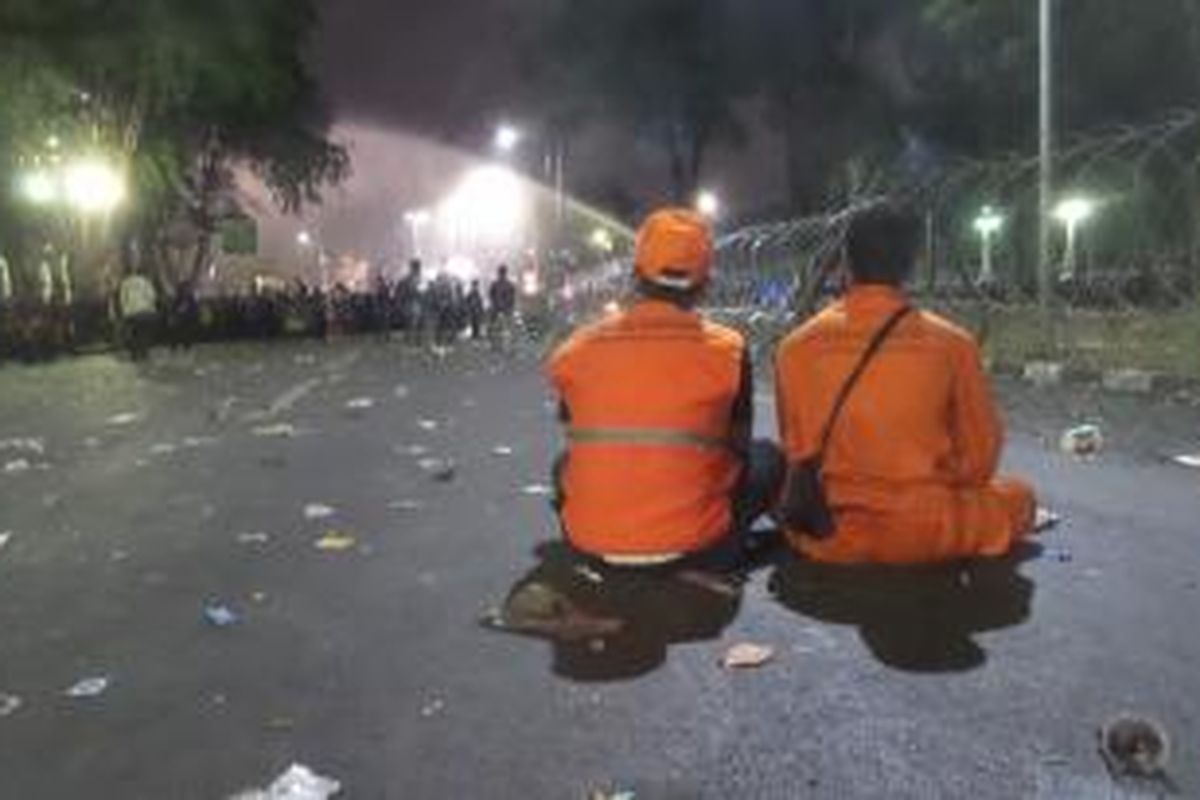 Dua petugas kebersihan dari Dinas Kebersihan DKI Jakarta terlihat sedang menyaksikan proses pembubaran paksa yang dilakukan polisi terhadap massa buruh dalam aksi unjuk rasa di seberang Istana Merdeka, Jakarta, Jumat (30/10/2015) malam.