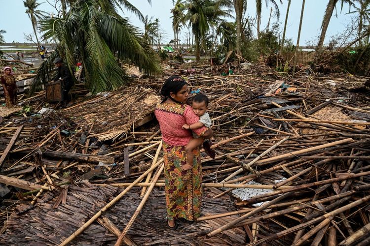 Seorang perempuan Rohingya menggendong bayinya di samping rumahnya yang hancur di kamp pengungsi Basara di Sittwe pada 16 Mei 2023, setelah Topan Mocha mendarat. Korban tewas akibat Topan Mocha yang melanda Myanmar telah mencapai 145 orang di negara itu, kata tim informasi junta dalam sebuah pernyataan pada 19 Mei.