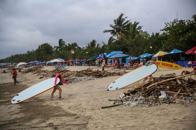 Wisatawan setelah melakukan surfing melintas di samping tumpukan sampah saat musim angin barat di pesisir pantai Kuta, Bali, Jumat (14/12/2018). Akibat terjadinya musim angin barat di wilayah perairan Bali berdampak pada banyaknya sampah yang hanyut terbawa arus laut sehingga menumpuk di pinggiran pantai kawasan wisata kuta.