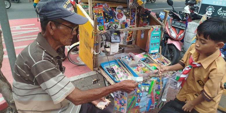 Ahmad melayani anak sekolah yang membeli mainan tradisional tembak bambu yang dijualnya di depan SDN Lateng Banyuwangi, Sabtu (11/1/2025).