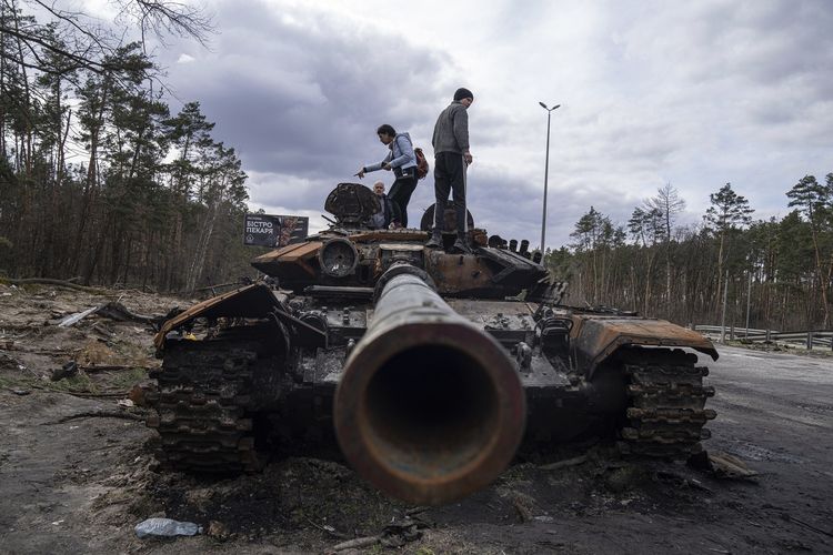 Warga lokal berdiri di atas tank Rusia yang hancur dalam pertempuran antara pasukan Rusia dan Ukraina di pinggiran Kyiv, Senin (11/4/2022).