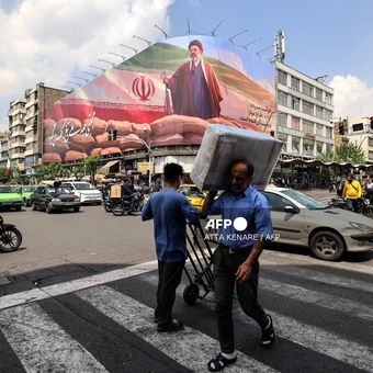 Seorang pria yang membawa bungkusan di pundaknya berjalan melewati papan reklame besar yang menampilkan Pemimpin Tertinggi Iran Ayatollah Mojtaba Khamenei di sebuah jalan di Teheran pada 20 April 2026.