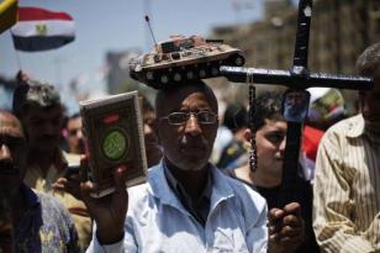 Pria Mesir mengenakan tank mainan di kepala sambil memegang salib dan salinan Al-Quran, saat berkumpul di Alun-alun Tahrir, setelah malamnya merayakan digulingkannya Presiden Mesir Muhammad Mursi, 4 Juli 2013.