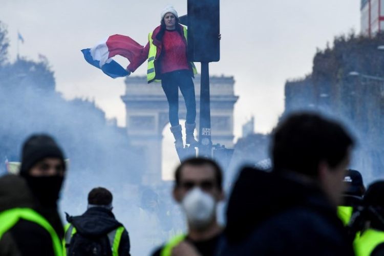 Seorang demonstran mengenakan rompi kuning memegang bendera Perancis saat berdiri di lampu merah di Champs Elysee, di Paris, Perancis, Sabtu (24/11/2018). Massa rompi kuning kembali berunjuk rasa untuk memprotes kenaikan harga minyak. (AFP/Bertrand Guay)