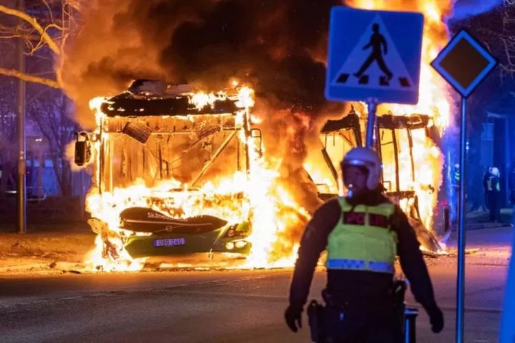 Seorang petugas polisi anti huru hara berdiri di samping bus kota yang terbakar di Kota Malmo, Sabtu malam 16 April 2022.