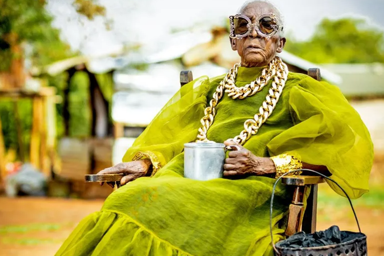 Nenek fashion icon, Margret Chola memegang cangkir logam penuh teh dengan mbaula yang menggantung di lengan kursi.