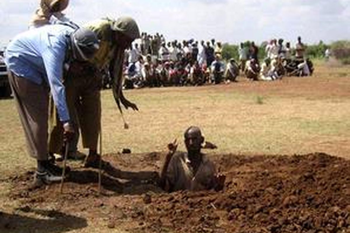 Saat-saat sebelum dieksekusi. Mohamed Abukar Ibrahim dikubur di tanah, keduan tangannya masih tampak.  Di latar belakang tampak warga desa yang dipaksa menonton proses eksekusi terhadap Ibrahim. 