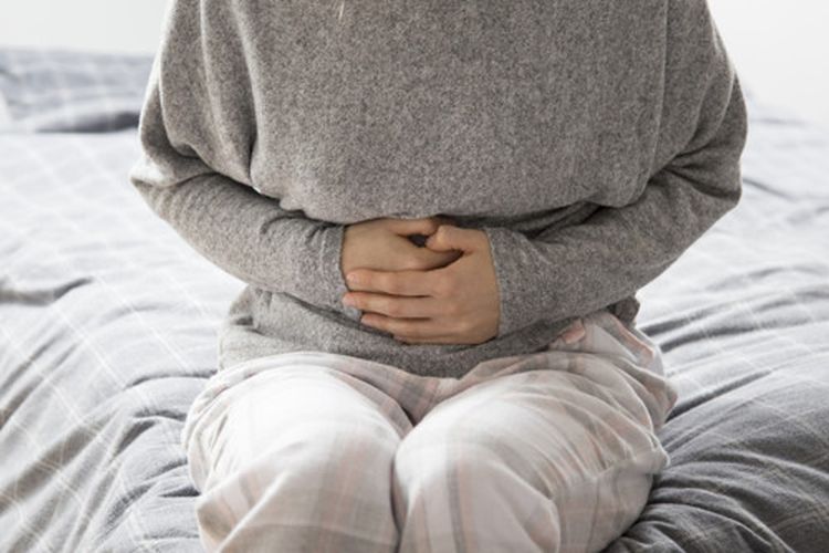 Sick woman in grey homewear sitting on bed, keeping hands on stomach, suffering from intense pain. Illness, stomach ache concept