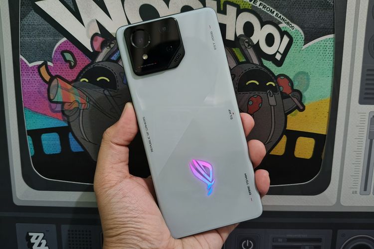 Desain belakang Asus ROG Phone 8. Tampilan punggung ponsel ini kini lebih sederhana dan minimalis. Bisa dilihat, lampu Aura RGB seakan menyatu dengan bodi ponsel yang dihiasi dengan berbagai kata ikonik ROG. Ada pula modul kamera belakang yang memuat tiga kamera, terdiri dari kamera utama 50 MP, ultrawide 13 MP, dan macro 5 MP.