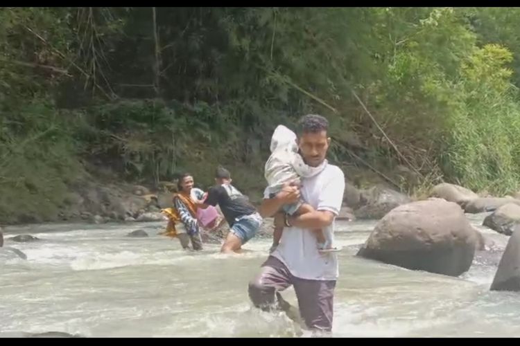 Elvis Jehama, anggota DPRD Manggarai Timur yang juga Sekretaris.Komisi C dan Ketua Fraksi Nasdem, memyeberangi sungai Waemokel sambil menggendong seorang anak dari satu ibu yang menyeberangi sungai itu, Sabtu (11/1/2025).