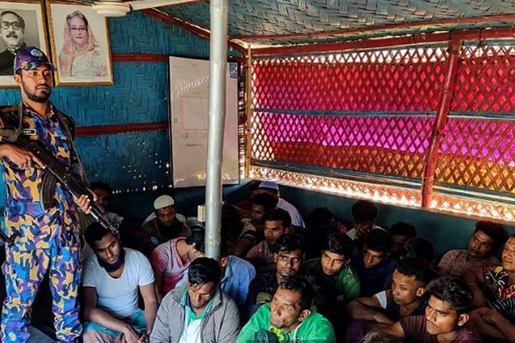 Foto selebaran yang diambil pada 28 Oktober 2022, dan dirilis oleh Batalyon Polisi Bersenjata Bangladesh (APBN) ini menunjukkan pengungsi Rohingya yang ditahan duduk di sebelah petugas keamanan setelah penumpasan di kamp pengungsi Rohingya di Ukhia 28, 2022.