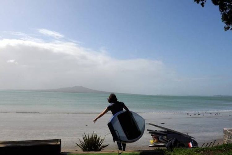 Seorang pria membawa sebuah papan selancar di Pantai Takapuna, sebelah utara Auckland, Selandia Baru. (AFP via Channel News Asia)