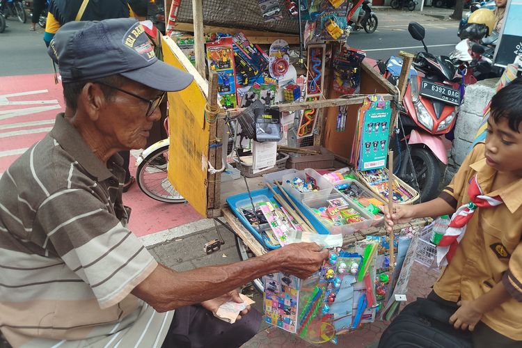Ahmad melayani anak sekolah yang membeli mainan tradisional tembak bambu yang dijualnya di depan SDN Lateng Banyuwangi, Sabtu (11/1/2025).