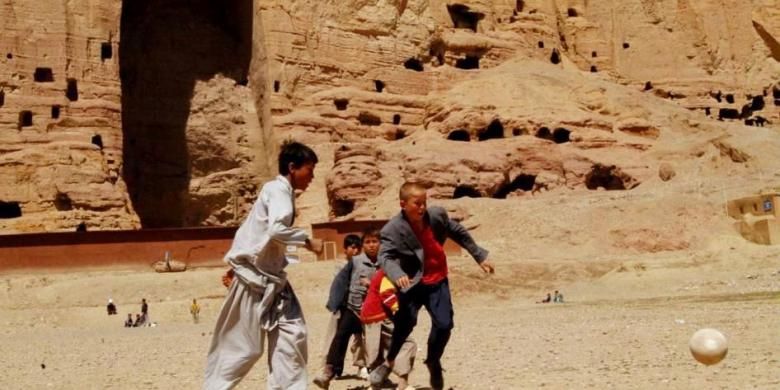 Anak laki-laki Afghanistan bermain sepak bola di depan area patung Buddha raksasa pernah berdiri di Provinsi Bamiyan, 9 April 2007. Patung-patung Buddha kuno dihancurkan oleh Taliban pada Maret 2000, meskipun ada protes internasional untuk menghentikan penghancuran.