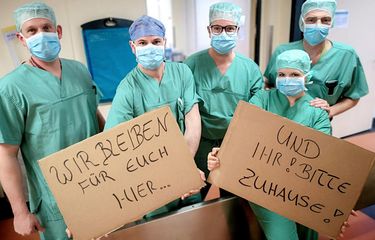 Sekelompok perawat di Jerman membawa tulisan di kardus, yang dalam bahasa Indonesia berarti Kami akan tetap di sini untuk kalian, dan kalian tolong tetap di rumah. Foto diambil pada 19 Maret 2020.