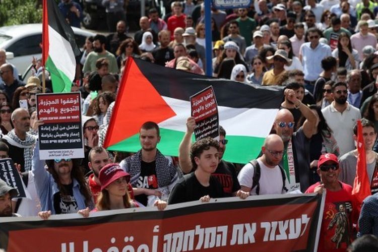 Orang-orang membentangkan spanduk dan mengibarkan bendera nasional Palestina saat unjuk rasa yang menandai peringatan ke-48 Hari Tanah, di kota Arab-Israel Deir Hanna di wilayah Galilea utara pada 30 Maret 2024. Hari Tanah memperingati peristiwa 30 Maret 1976, ketika pasukan Israel menembak dan menewaskan enam orang di antara warga Arab Israel dan Palestina yang memprotes penyitaan tanah. 