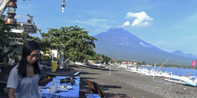 Pramusaji menyiapkan makanan untuk wisatawan saat asap putih terembus dari kawah Gunung Agung terlihat dari Amed, Karangasem, Bali, Kamis (7/12/2017). Gunung Agung yang berada dalam status level IV atau awas kembali mengembuskan asap putih tebal dari kawahnya.  