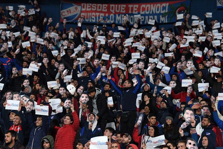 Para pendukung Nacional mengenakan topeng Luis Suarez dan memegang poster bertuliskan #SuarezANacional dalam laga lanjutan Torneo Intermedia, divisi teratas Liga Uruguay, kontra Cerrito, Kamis (21/7/2022) waktu setempat. Aksi ini dilakukan sebagai bentuk dukungan agar Luis Suarez kembali ke Nacional.