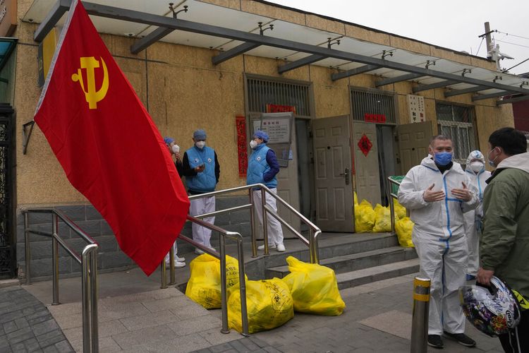 Pekerja komunitas di luar komunitas yang terkunci berbincang di dekat bendera Partai Komunis dan kantong sampah berlabel limbah berbahaya pada Kamis, 17 Maret 2022, di Beijing.