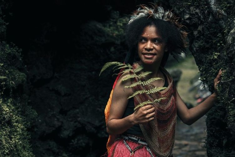 Closeup gadis muda eksotis suku dani papua dengan pakaian tradisional tersenyum di tengah hutan.