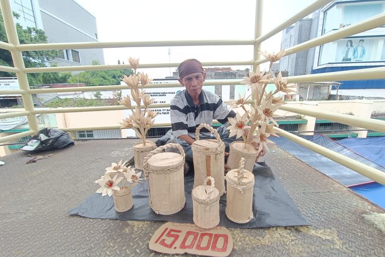 Ridwan (50) dengan dagangannya, yakni kerajinan tangan berupa dekorasi bunga dan celengan dari pelepah pisang, di Jatinegara, Jakarta Timur, Kamis (28/3/2024).
