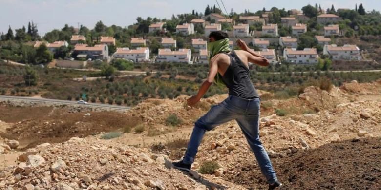Seorang pengunjuk rasa melemparkan batu ke arah tentara Israel selama bentrokan menyusul perlawanan atas perampasan tanah Palestina untuk memperluas pemukiman Yahudi Hallamish (latar belakang), Jumat (28/8/2015) di Desa Nabi Saleh, Tepi Barat, dekat Ramallah.