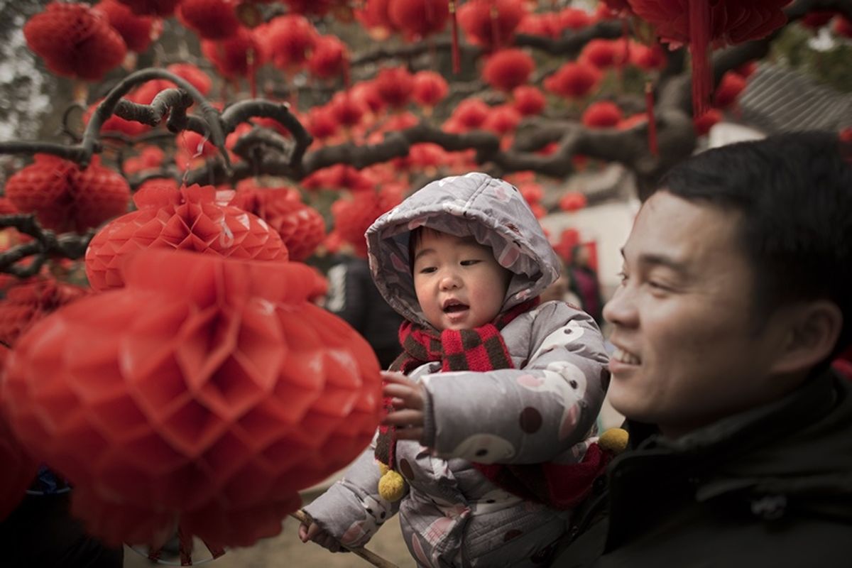 Seorang pria menggendong anak di samping pohon dihiasi lentera yang dipasang untuk menyambut Imlek, di taman Ditan, Beijing, China, Sabtu (17/2/2018). (AFP/Nicolas Asfouri)