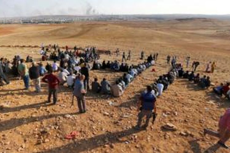 Para pengungsi Kurdi berkumpul di sebuah bukit di kota Mursipitnar, Turki untuk menyaksikan perkembangan pertempuran di kota Kobani yang terletak tak jauh dari perbatasan Turki-Suriah. Di kejauhan nampak asap mengepul akibat pertempuran di kota Kobani.