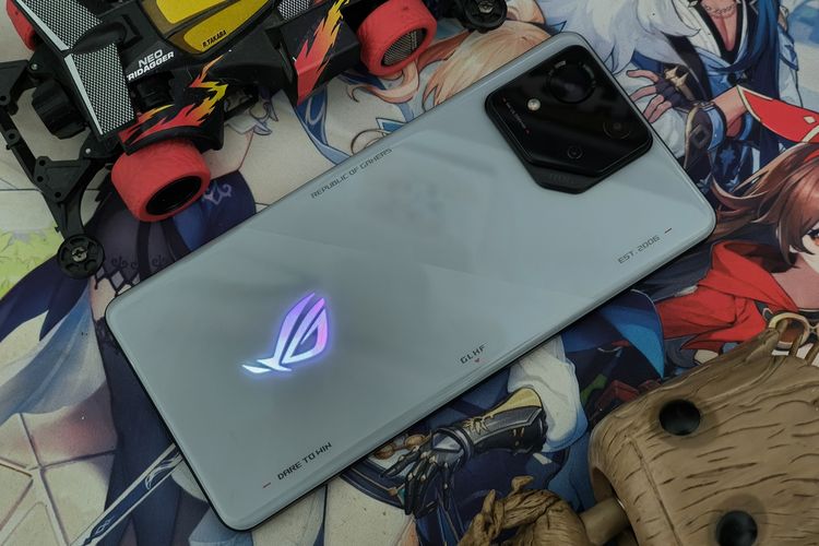 Desain belakang Asus ROG Phone 8. Tampilan punggung ponsel ini kini lebih sederhana dan minimalis. Bisa dilihat, lampu Aura RGB seakan menyatu dengan bodi ponsel yang dihiasi dengan berbagai kata ikonik ROG. Ada pula modul kamera belakang yang memuat tiga kamera, terdiri dari kamera utama 50 MP, ultrawide 13 MP, dan macro 5 MP.