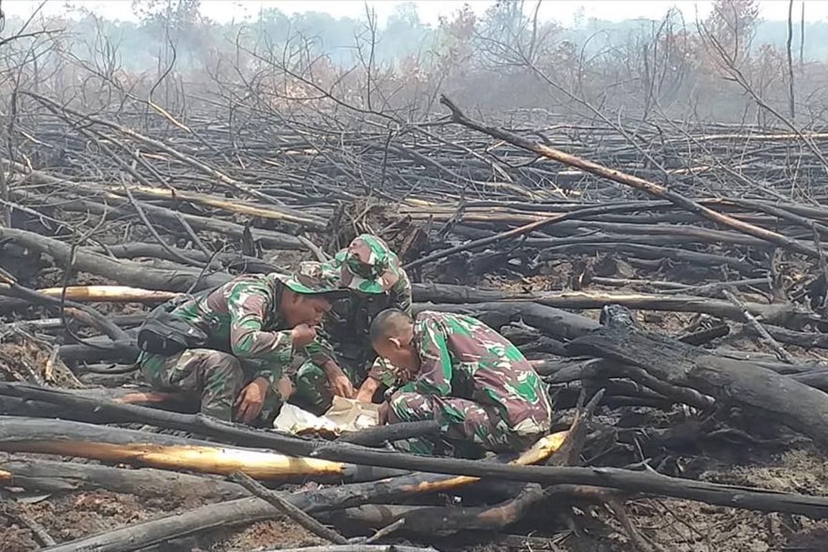 Serda Imam Effendi (tengah) dan dua prajurit TNI AD lainnya sedang makan siang di tumpukan kayu bekas terbakar di lahan yang sudah mereka padamkan di Desa Bedagu, Kecamatan Langgam, Kabupaten Pelalawan, Riau, Sabtu (3/8/2019).