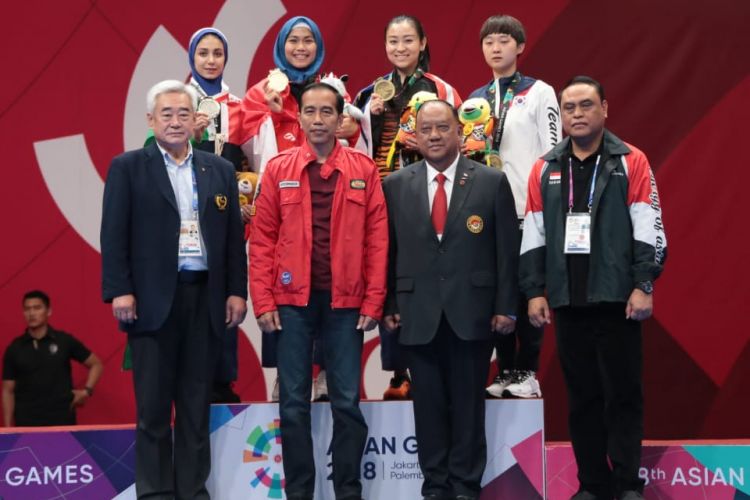 Defia Rosmaniar berfoto di belakang Presiden Republik Indonesia, Joko Widodo, bersama Chef de Mission (CdM) Tim Indonesia, Komjen Pol. Syafruddin, seusai pengalungan medali emas taekwondo di JCC Plenary Hall, 19 Agustus 2018.