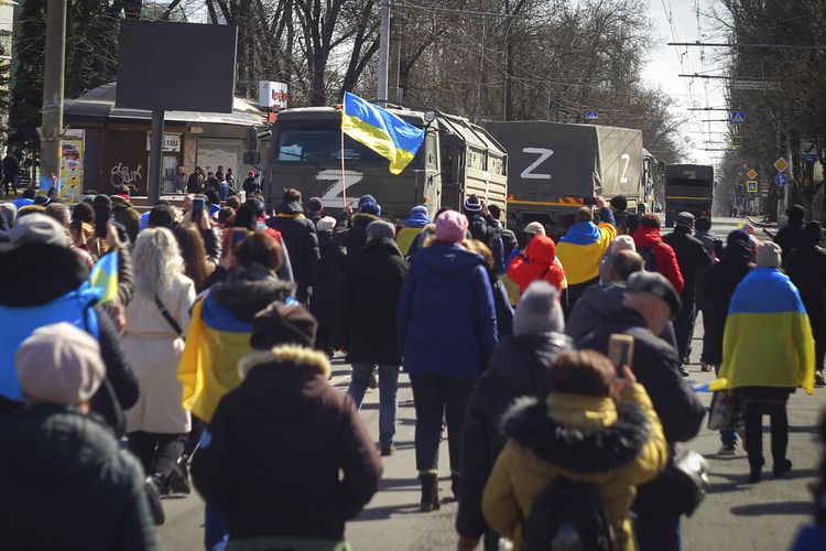 Orang-orang membawa bendera Ukraina berjalan menuju truk tentara Rusia saat unjuk rasa menentang pendudukan Rusia di Kherson, Ukraina, Minggu (20/3/2022). PBB mengecam intimidasi yang dilakukan Rusia terhadap warga yang menentang perang Rusia.
