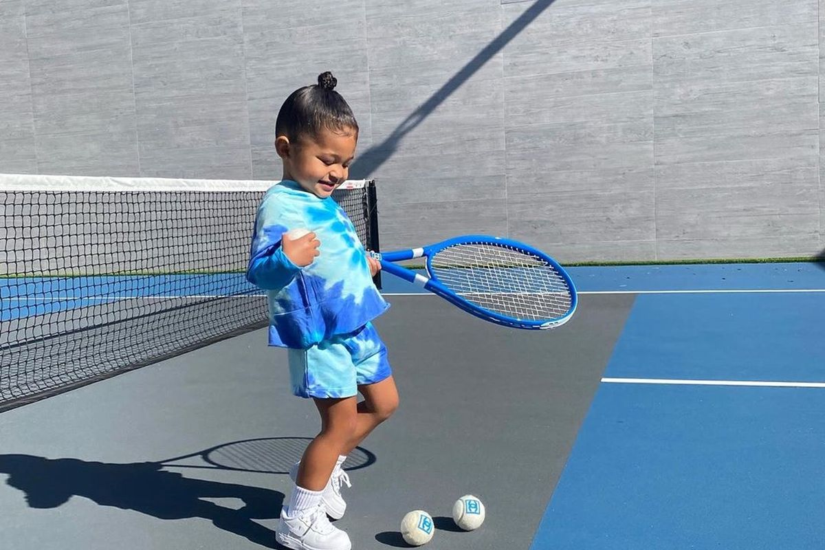 Putri Kylie Jenner, Stormi Webster, mengenakan pakaian serba biru sambil memegang raket tenis Chanel di tangan kirinya.