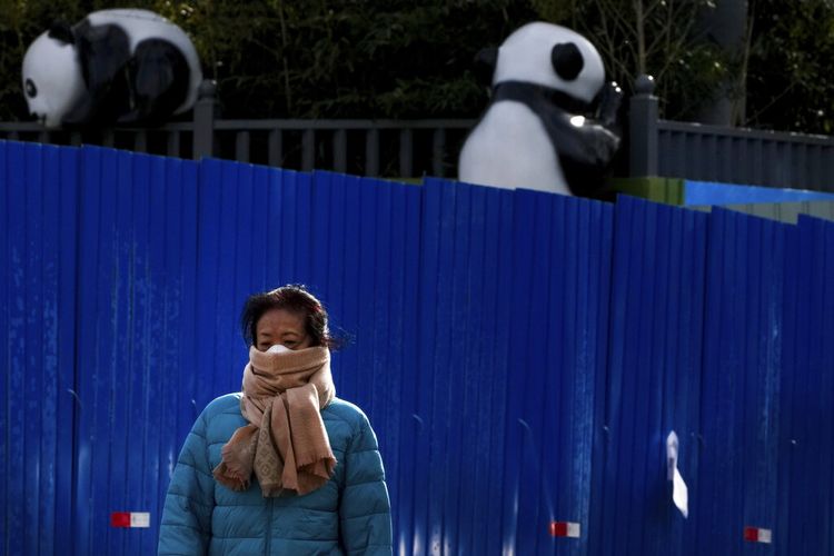 Seorang wanita yang mengenakan masker berjalan melewati penghalang logam yang dipasang di depan patung panda di Beijing, Selasa, 29 November 2022.