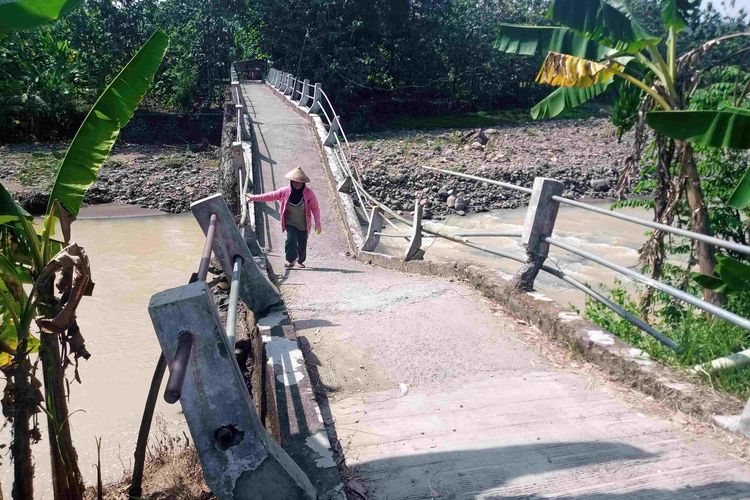 Seorang warga melewati jembataan Dusun Banaran yang ambles diterjang banjir.