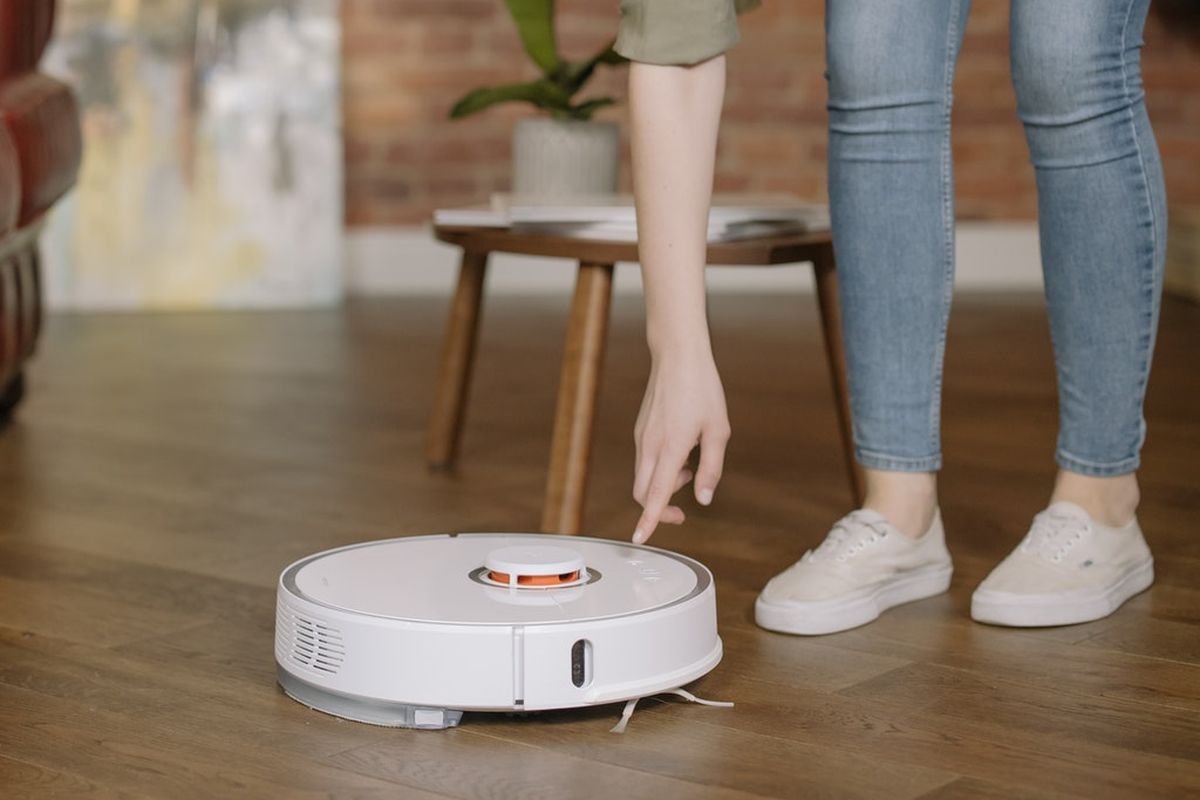 Seorang wanita menyalakan robot pembersih lantai otomatis berwarna putih di ruang tamu dengan lantai kayu