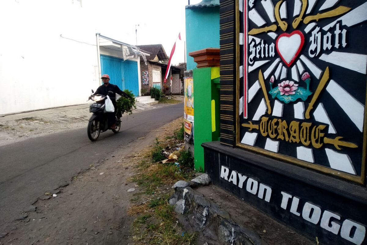 Seorang pria mengangkut rumput pakan ternak melewati depan tugu perguruan silat PSHT di Desa Tlogo, Kecamatan Kanigoro, Kabupaten Blitar, Selasa (15/8/2023).