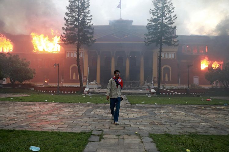Seorang pria mengenakan bendera Nepal berjalan di depan kantor presiden yang dibakar di Kathmandu, Nepal, Selasa (9/9/2025). Demo Nepal pecah sejak Jumat (5/9/2025) setelah pemerintah memblokir 26 media sosial, dan amarah publik atas korupsi pejabat.