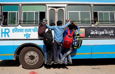 Para pekerja migran bergantungan ke sebuah pintu bus yang bergerak saat mereka kembali ke desa mereka, saat diberlakukan lockdown nasional selama 21 hari untuk membatasi penyebaran penyakit virus corona (COVID-19) di Ghaziabad, pinggiran kota New Delhi, India, Minggu (29/3/2020).