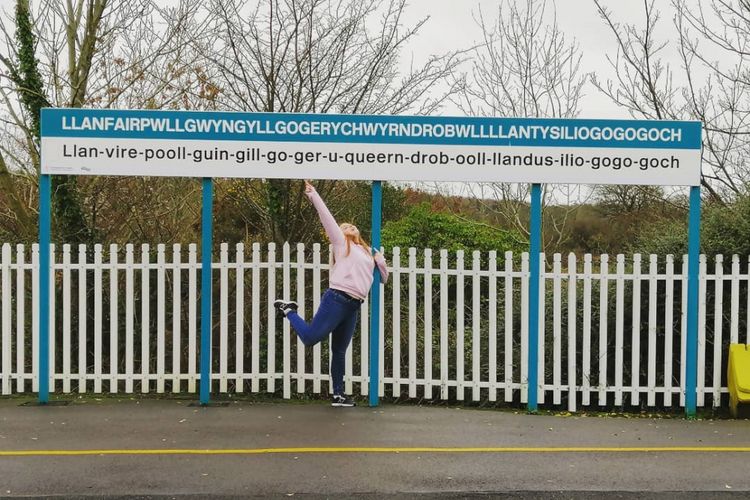 Seseorang berpose di depan papan nama desa dengan nama terpanjang di Inggris dan Eropa.